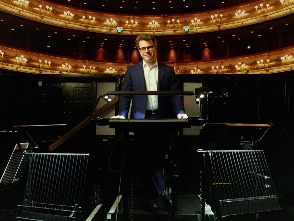 Jakub Hrůša, wearing a dark suit and glasses, stands behind a conductor's podium in the orchestra pit at the Royal Opera House. An ornate balcony and chandeliers are visible in the background as he smiles at the camera.