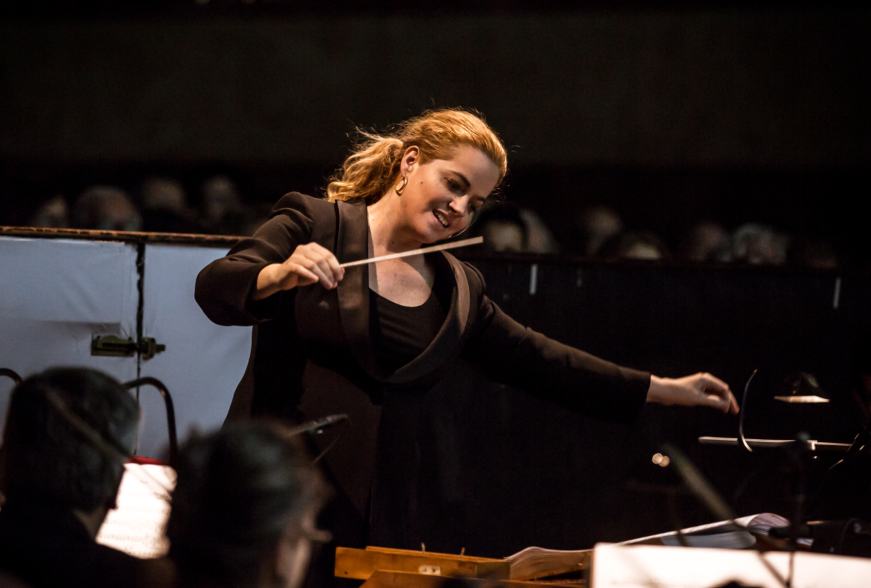 Speranza Scappucci, a female conductor leads an orchestra, holding a baton in her right hand and gesturing expressively with her left. Her hair is tied back in a ponytail, and she wears a black blazer over a dark top. Blurred outlines of darkened seating fill the background.