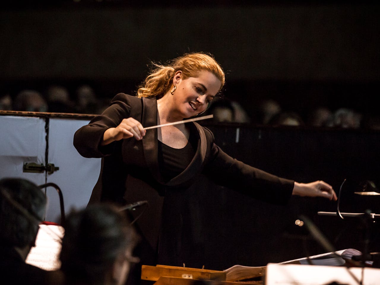 Speranza Scappucci, a female conductor leads an orchestra, holding a baton in her right hand and gesturing expressively with her left. Her hair is tied back in a ponytail, and she wears a black blazer over a dark top. Blurred outlines of darkened seating fill the background.