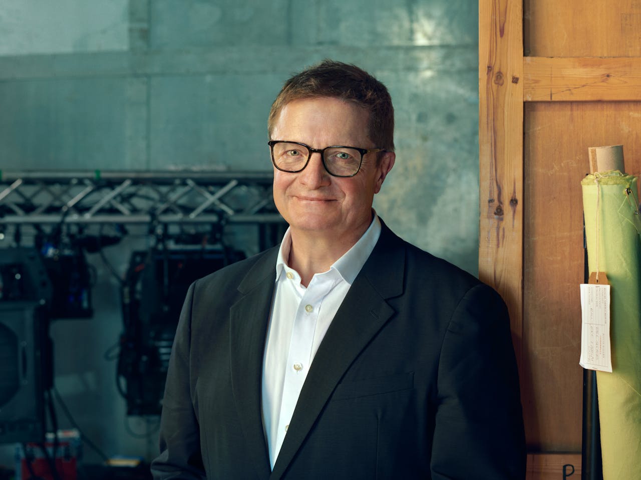 Alex Beard, wearing glasses and a navy blazer stands in front of a muted green wall, smiling slightly. He has short brown hair and is dressed in a white shirt. In the background, there is professional lighting rig, and rolls of coloured materials leant against a wooden crate.