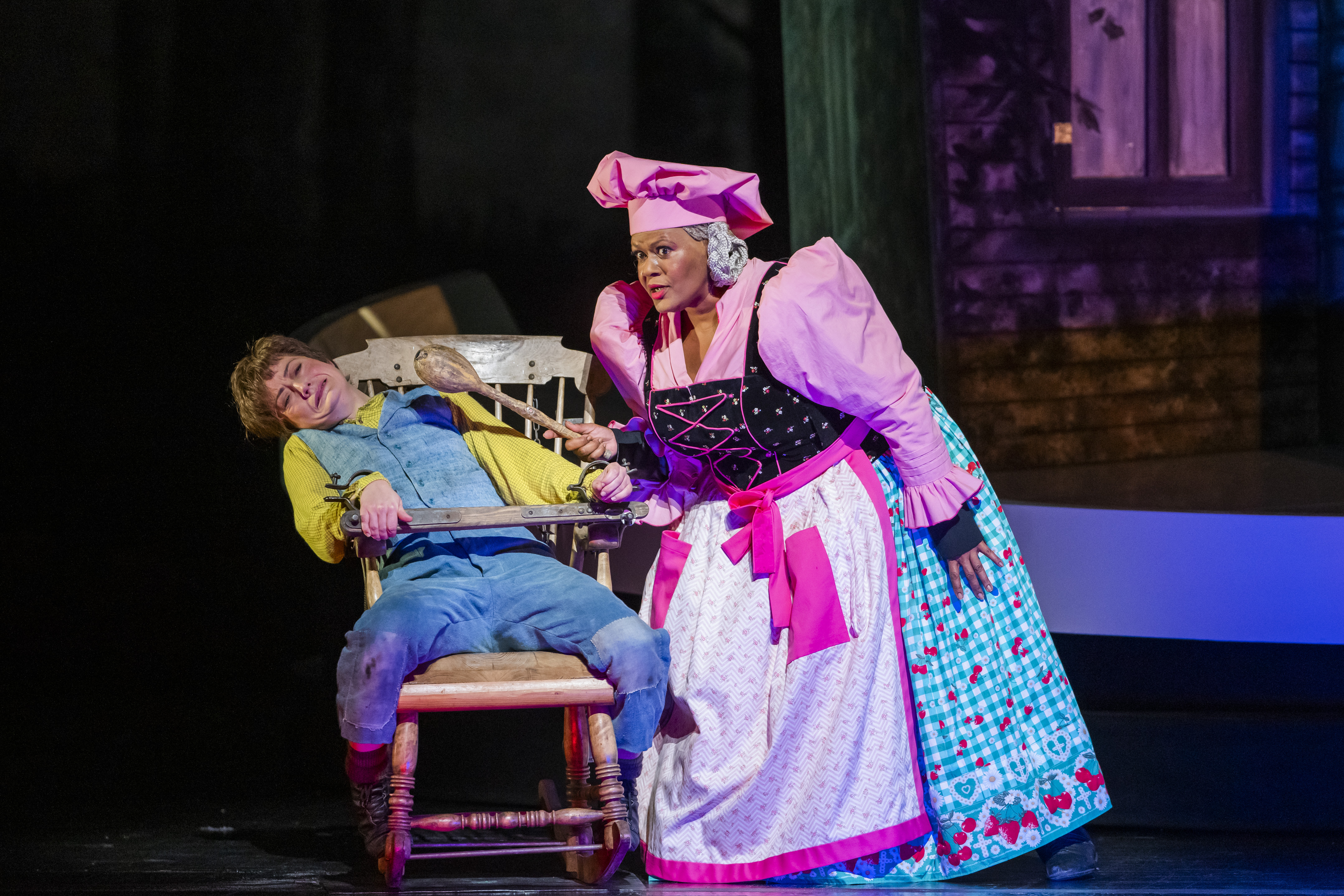 A woman dressed in a yellow shirt and blue overalls sits in a chair. They are recoiling from a woman dressed as a chef, holding a large wooden spoon.