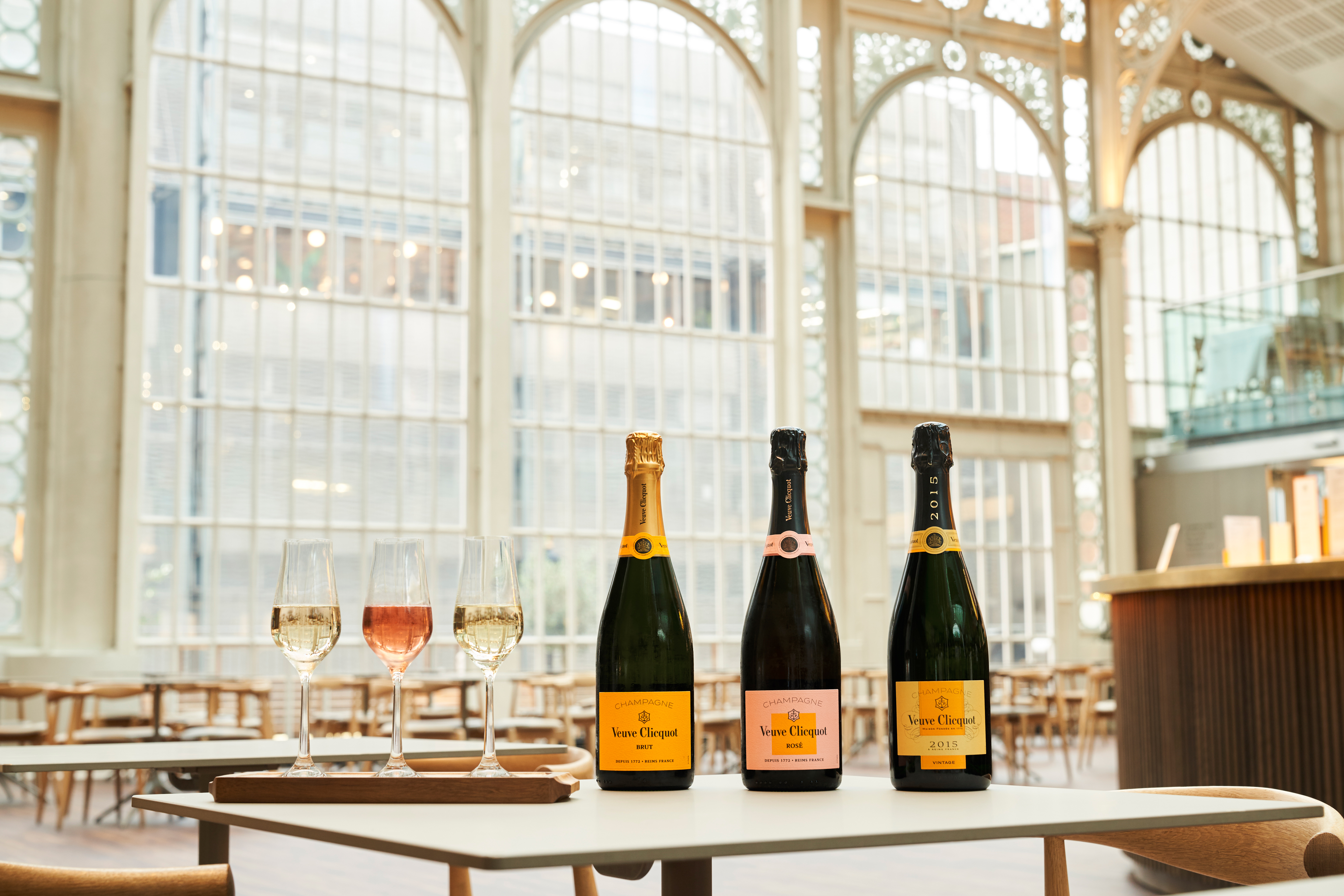 Three bottles of Veuve Clicquot champagne sit on a table with three filled glasses either side. The table sits in the middle of a large ornate window which is part of the Paul Hamlyn Hall in the Royal Opera House.