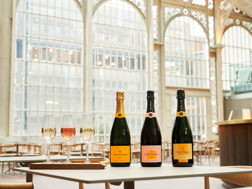 Three bottles of Veuve Clicquot champagne sit on a table with three filled glasses either side. The table sits in the middle of a large ornate window which is part of the Paul Hamlyn Hall in the Royal Opera House.