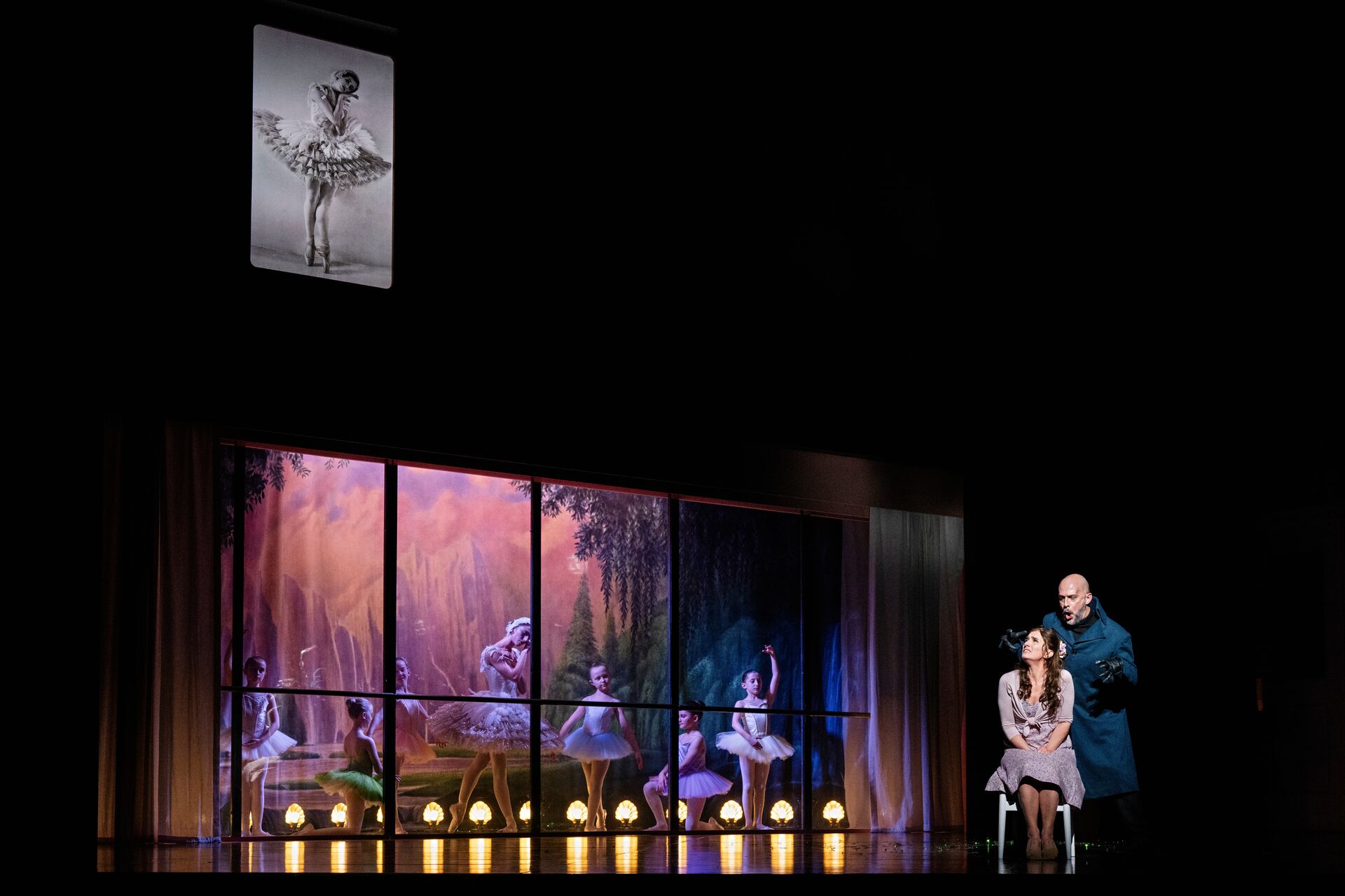 A man in dark clothes stands behind a frightened woman sitting on a chair. There are bright purple windows lit up around with ballet dancers behind them.