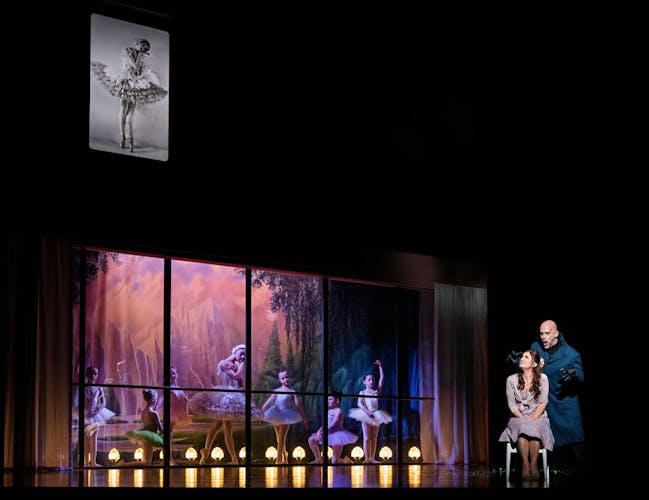 A man in dark clothes stands behind a frightened woman sitting on a chair. There are bright purple windows lit up around with ballet dancers behind them.