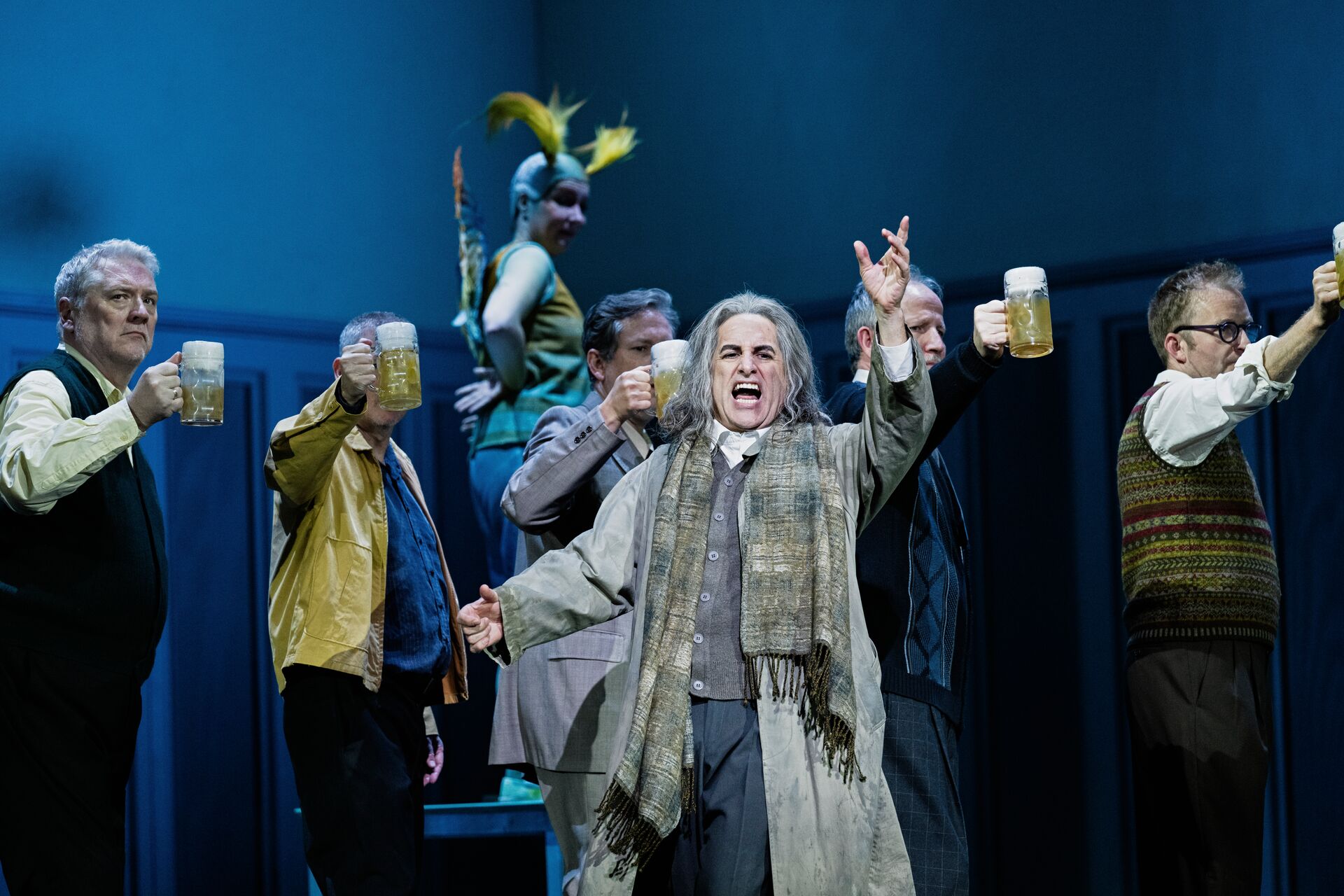 A singer dressed as an old man stands in the foreground. Behind up are several men holding up beers.