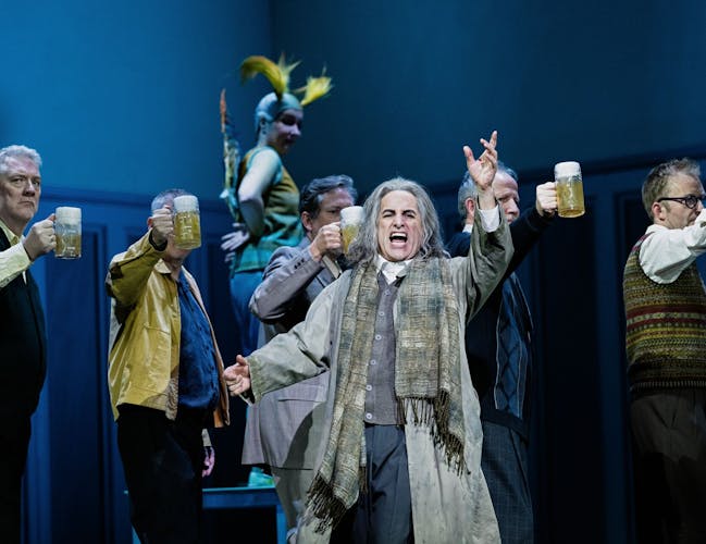 A singer dressed as an old man stands in the foreground. Behind up are several men holding up beers.