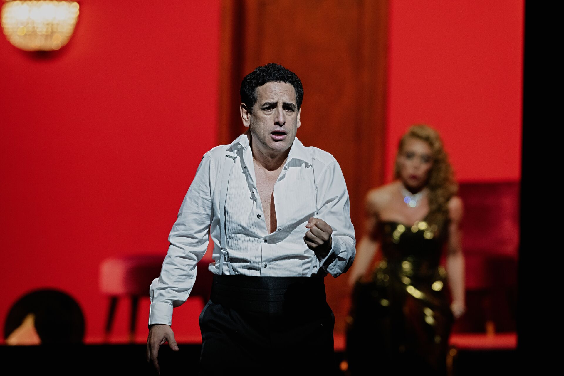 A man in an open white shirt stands in the foreground. He looks distressed. In the background a blond woman wearing a gold dress stands against a red wall.