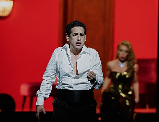 A man in an open white shirt stands in the foreground. He looks distressed. In the background a blond woman wearing a gold dress stands against a red wall.