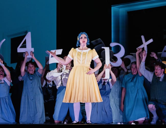 A group of school children hold up large numbers. In the foreground a singer plays the role of a doll. She is wearing a yellow dress and has blue hair.