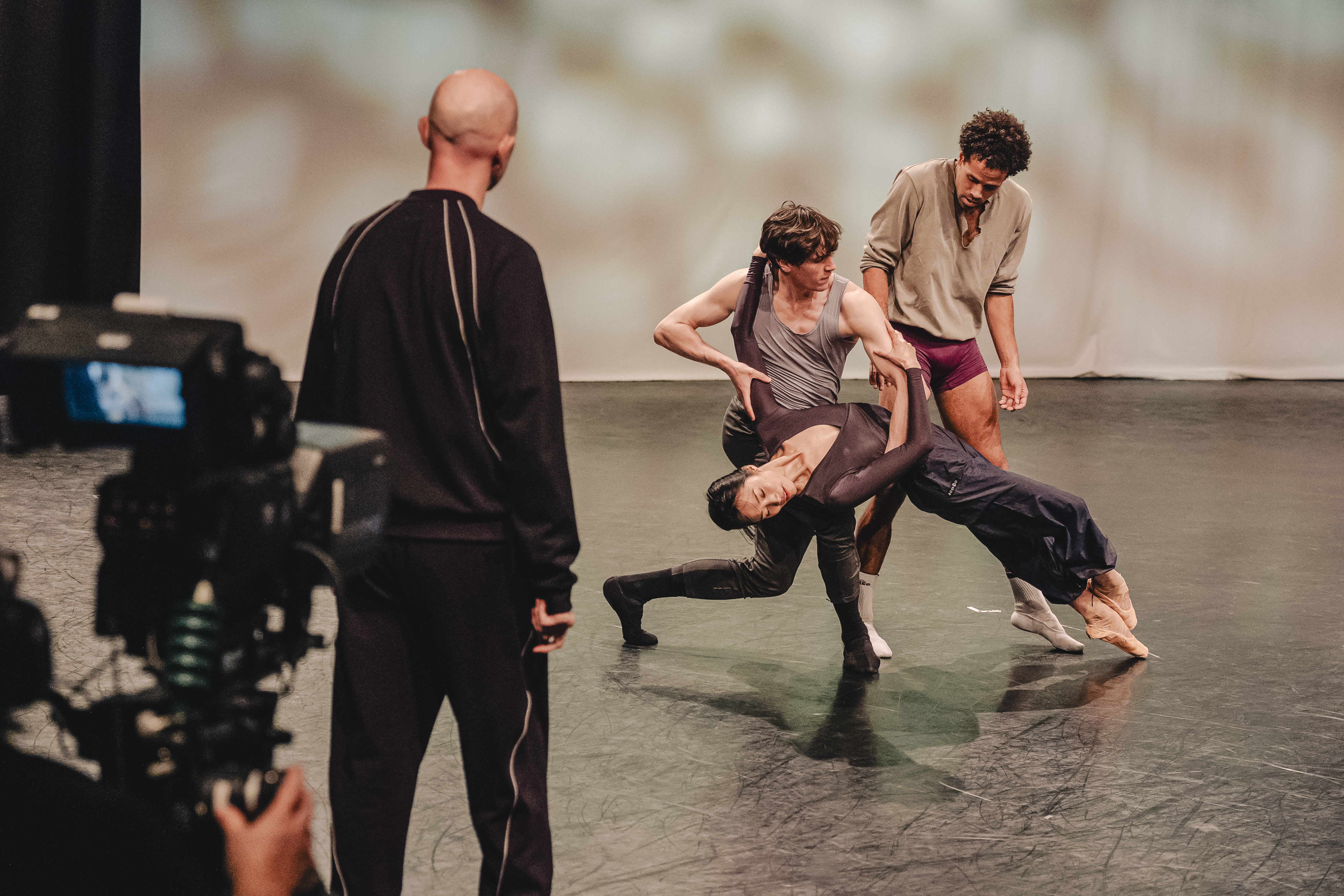 A film camera in the corner overlooks the back of Resident Choreographer Sir Wayne Mcgregor as he watches three dancers of The Royal Ballet rehearse in the centre of a studio. One dancer is leaning against another and the third is performing behind them. 