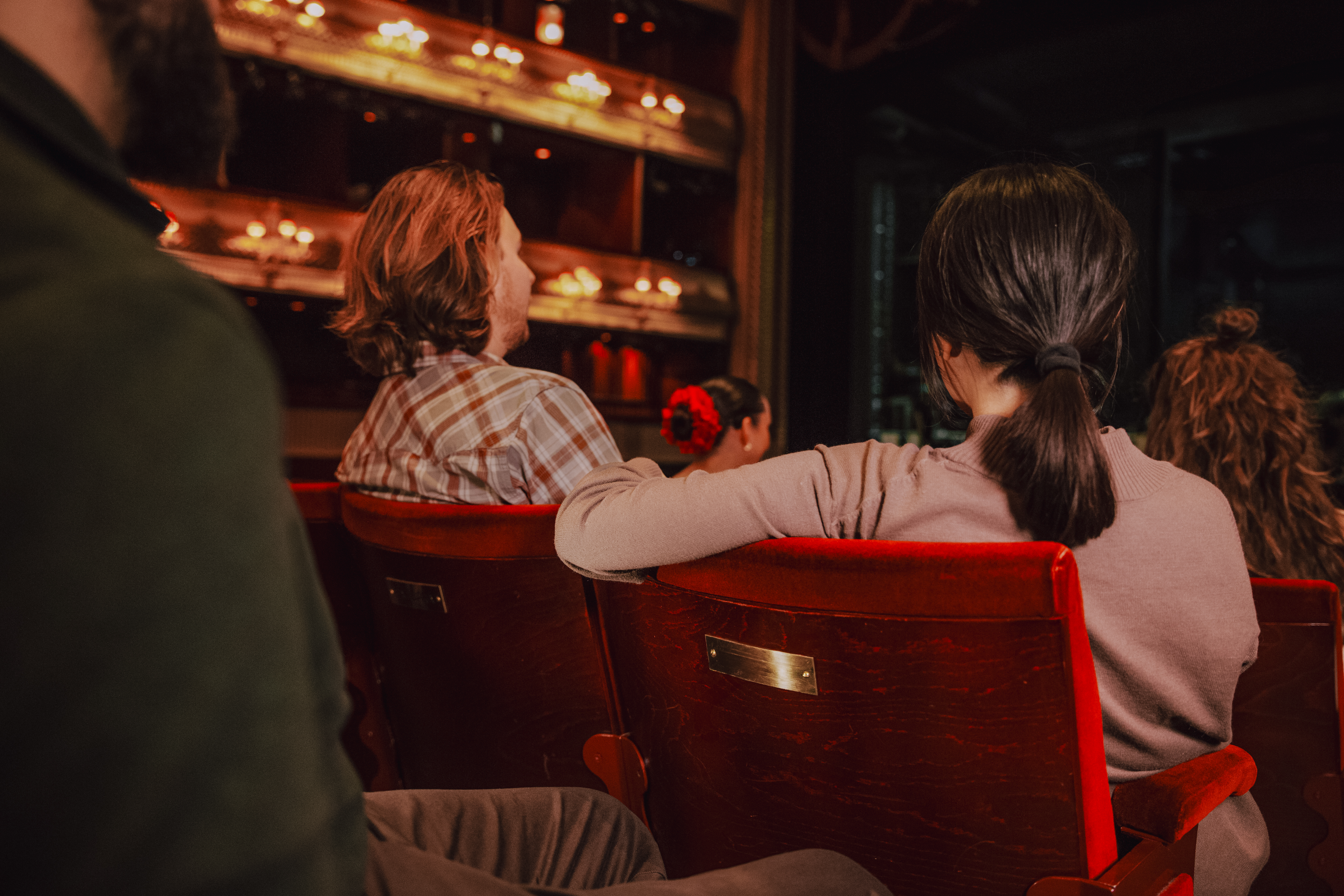 People sit on red velvet chairs in a theatre auditorium. On the back of the chair in the foreground is a gold plaque.