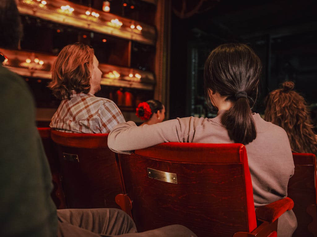 People sit on red velvet chairs in a theatre auditorium. On the back of the chair in the foreground is a gold plaque.