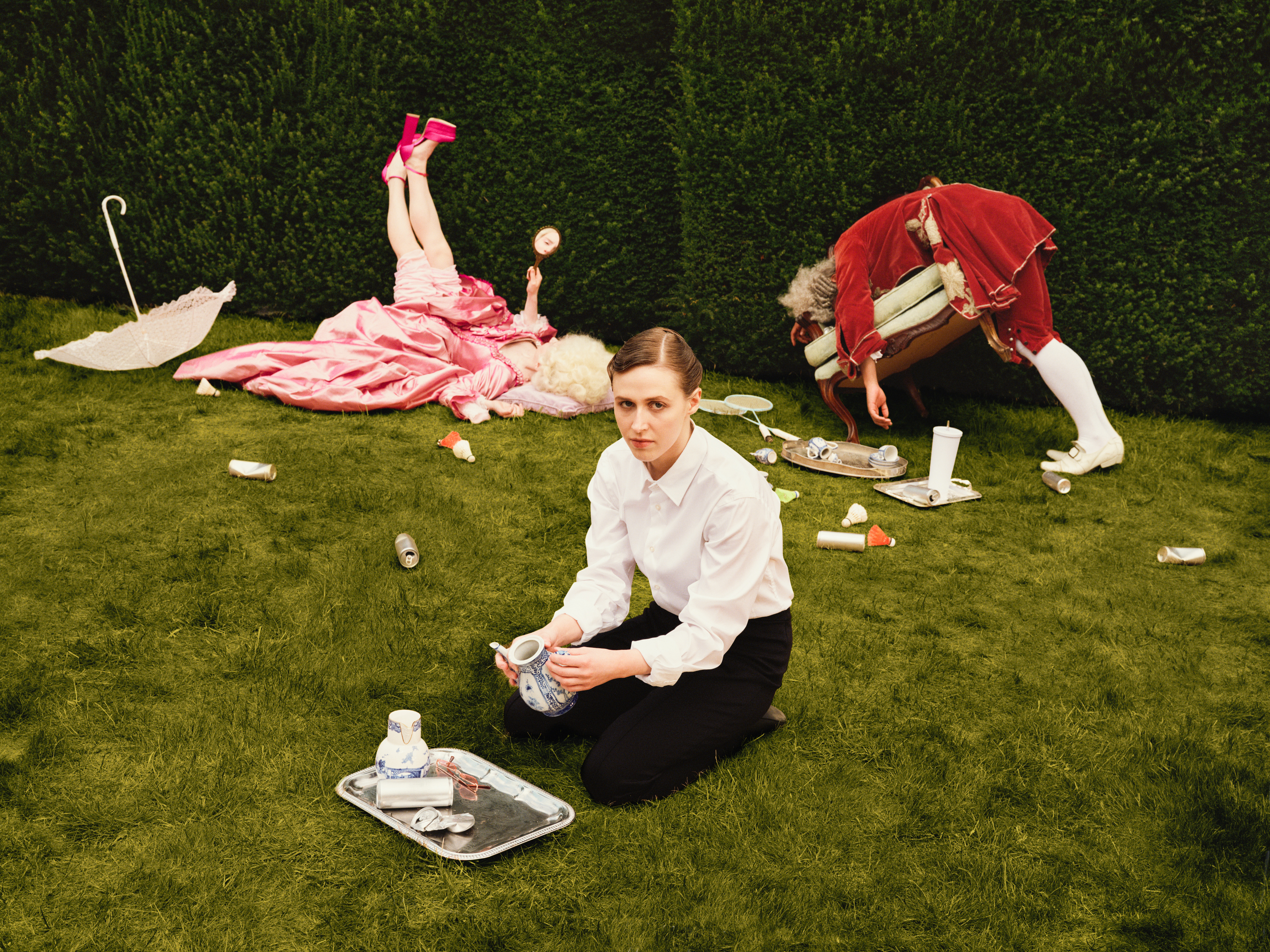 A woman in a maid costume, representing Despina in Netia Jones’s Così fan tutte, kneels on a lawn clearing out tea cups from the ground, surrounded by trays and scattered crockery. Behind her, two performers in elaborate pink and red period costumes lie and bend over among the garden furniture, creating a playful, chaotic scene. (3 of 4)