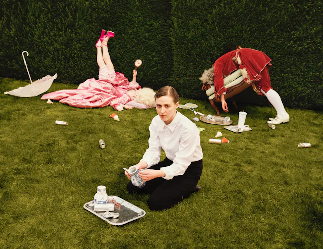 A woman in a maid costume, representing Despina in Netia Jones’s Così fan tutte, kneels on a lawn clearing out tea cups from the ground, surrounded by trays and scattered crockery. Behind her, two performers in elaborate pink and red period costumes lie and bend over among the garden furniture, creating a playful, chaotic scene. (3 of 4)