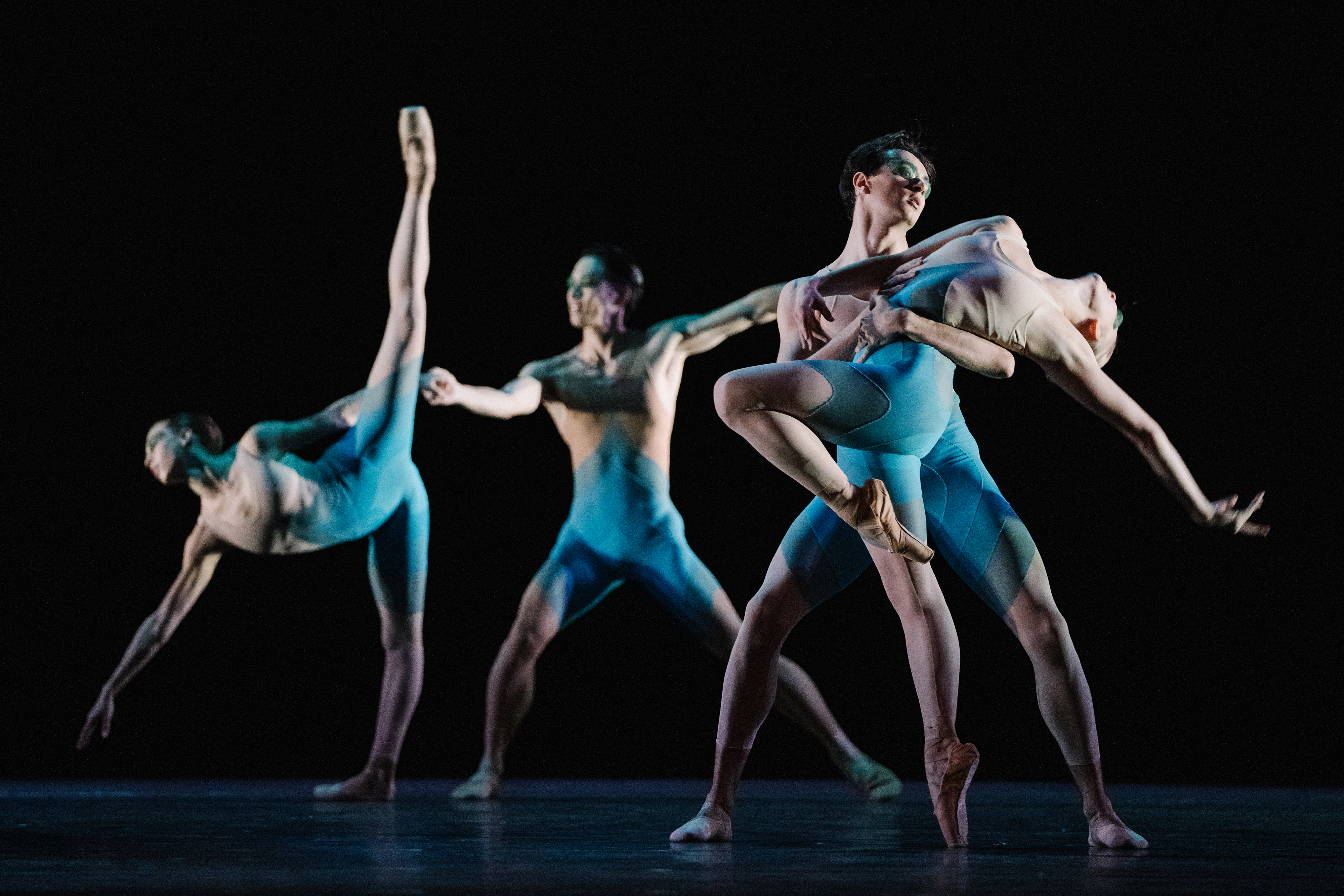 Four dancers of The Royal Ballet stand in different poses wearing blue leotards - one leans forward on one leg with one arm facing downward and their other leg raised high making an A shape, another lunges forward. The other two dancers hold one another.

