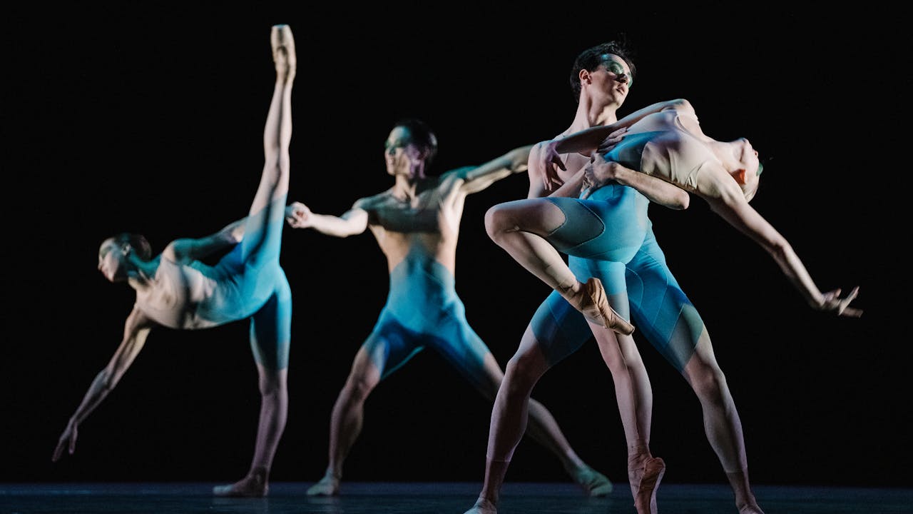 Four dancers of The Royal Ballet stand in different poses wearing blue leotards - one leans forward on one leg with one arm facing downward and their other leg raised high making an A shape, another lunges forward. The other two dancers hold one another.