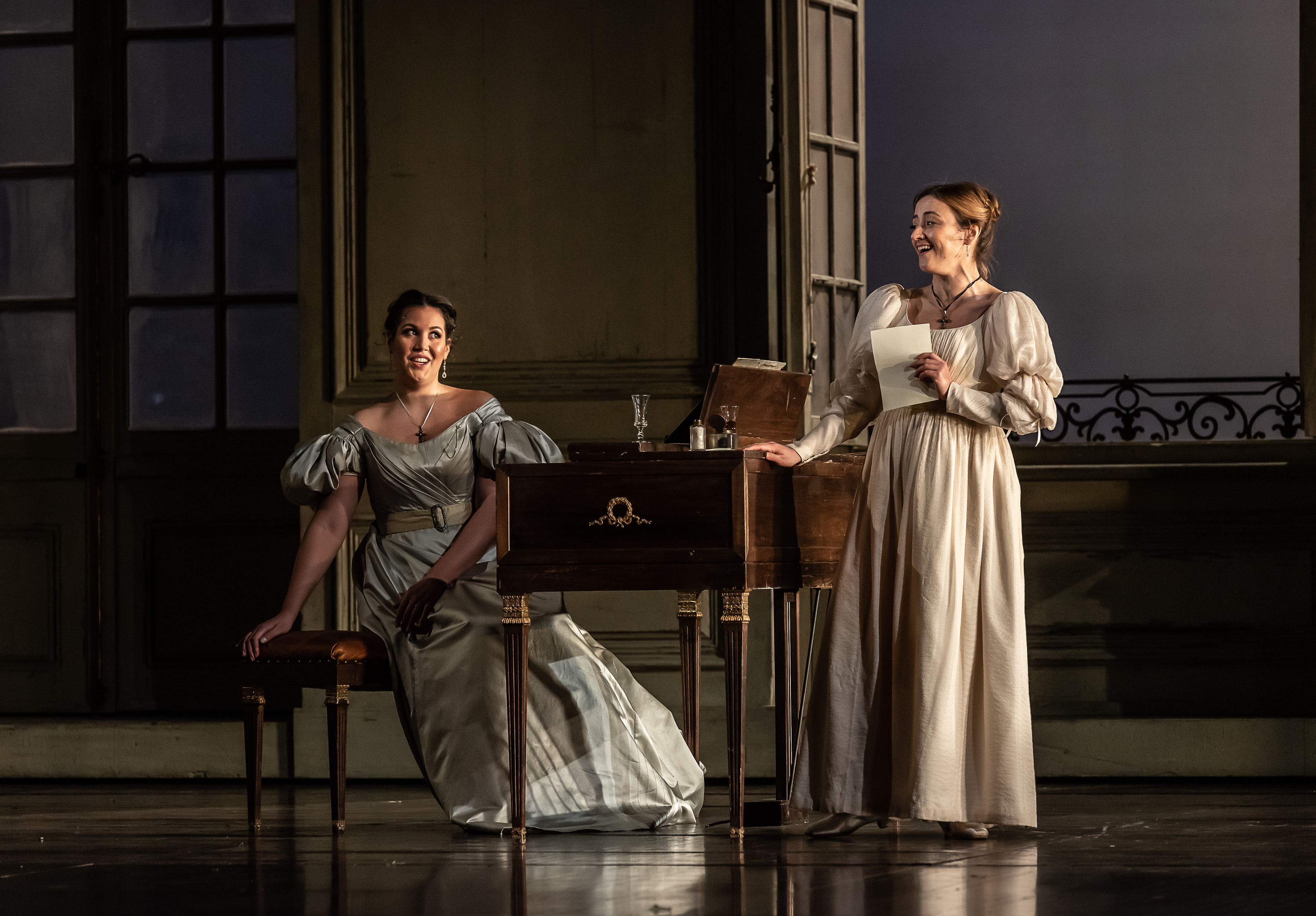 Two performers wearing long ballgowns are singing onstage at the Royal Opera House in a production of The Marriage of Figaro. One stands holding a piece of paper up to their chest. The other is sitting on a chair at a desk looking towards the other performer.