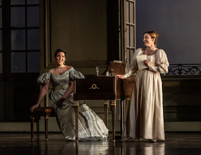 Two performers wearing long ballgowns are singing onstage at the Royal Opera House in a production of The Marriage of Figaro. One stands holding a piece of paper up to their chest. The other is sitting on a chair at a desk looking towards the other performer.