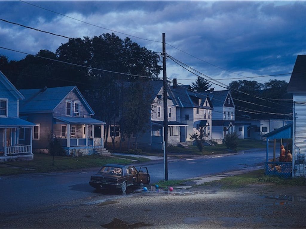 A suburban street at twilight with a telegraph pole and a car in the foreground.