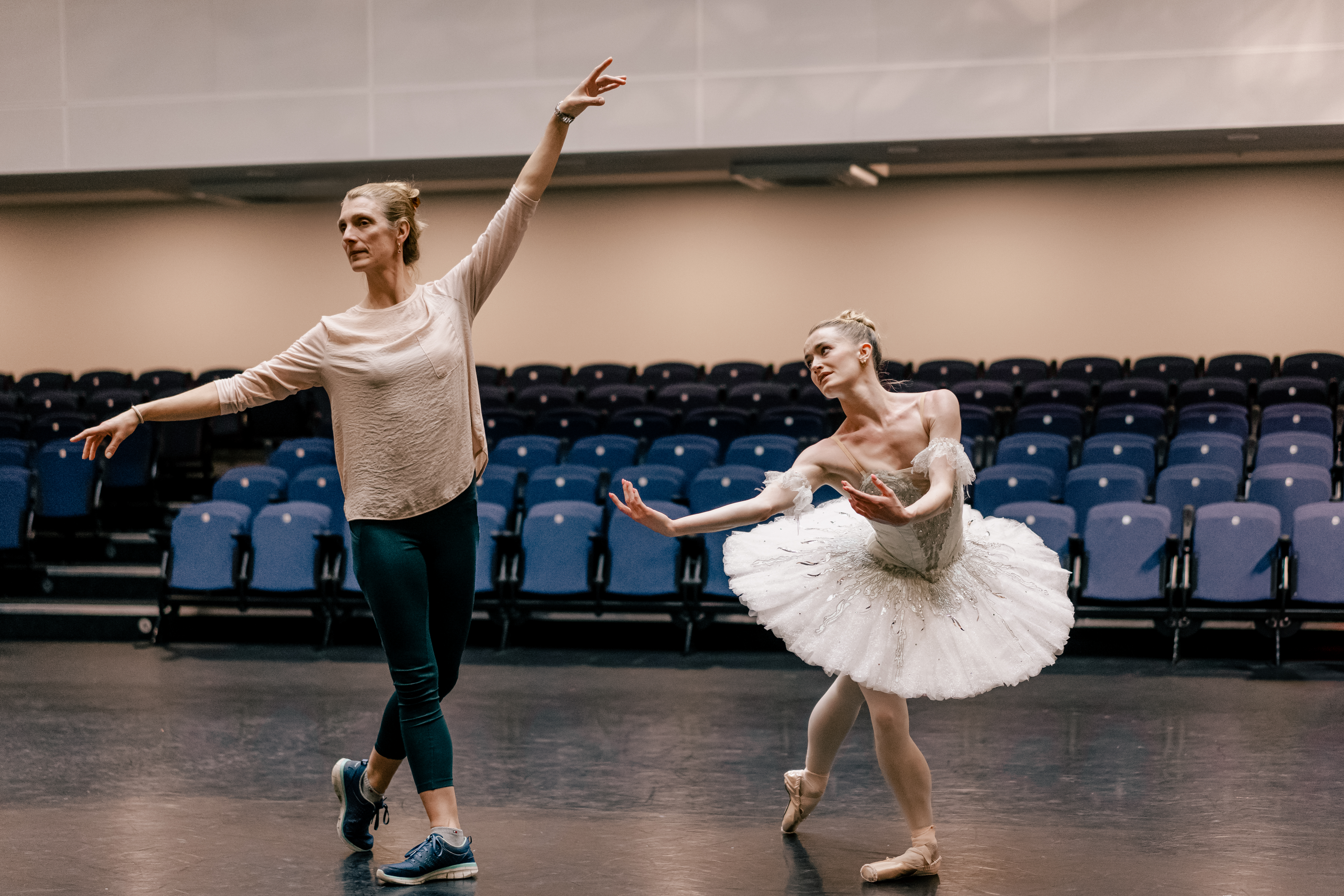 Zenaida Yanowsky, a répétiteur, demonstrates arm placement to dancer Anna Rose O'Sullivan, who is dressed in a white tutu. They are in a brightly lit rehearsal studio with a dark floor; blue raked seating is visible in the background behind them.