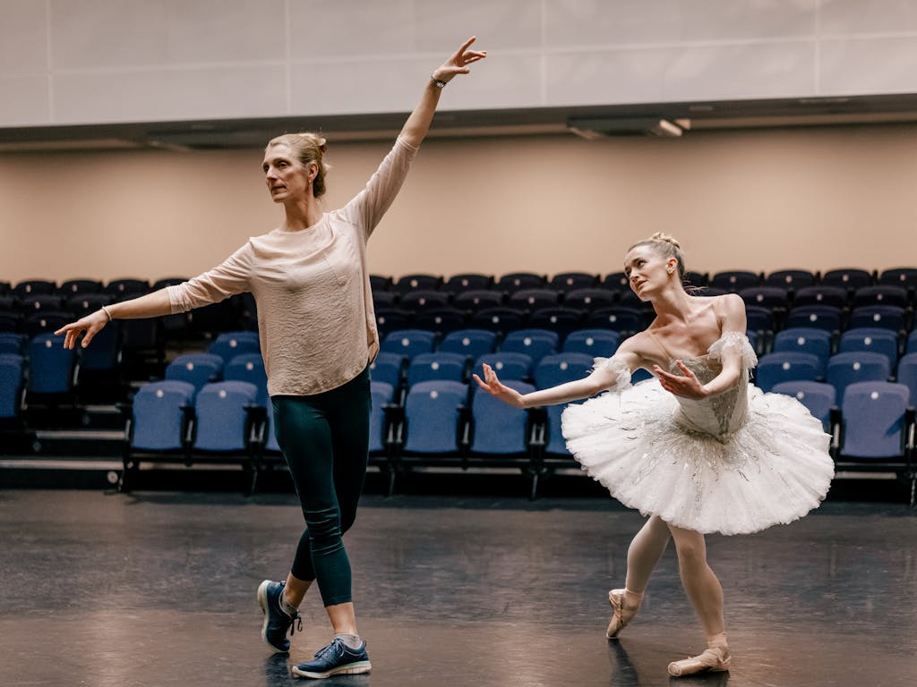 Zenaida Yanowsky, a répétiteur, demonstrates arm placement to dancer Anna Rose O'Sullivan, who is dressed in a white tutu. They are in a brightly lit rehearsal studio with a dark floor; blue raked seating is visible in the background behind them.