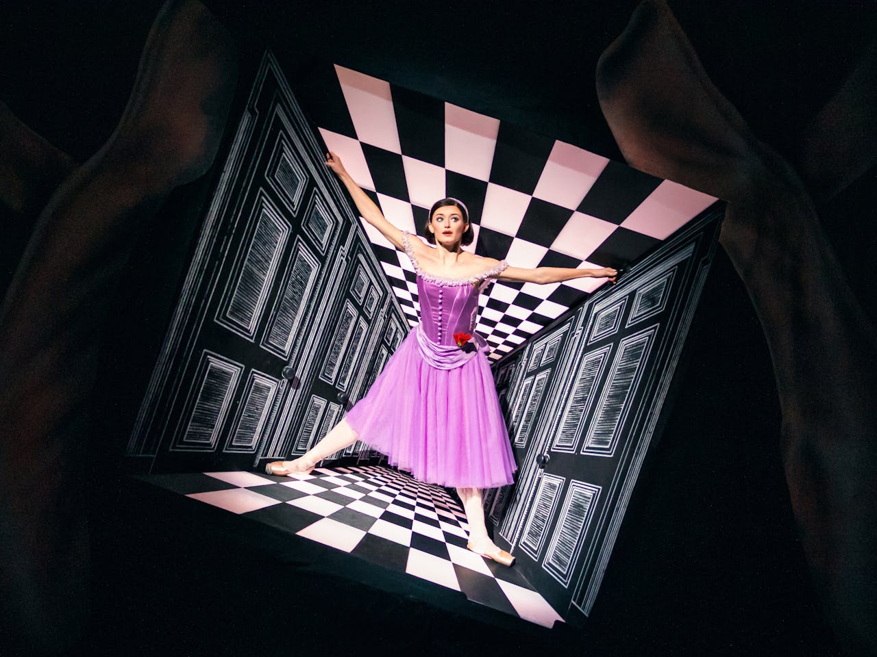 Anna Rose O’Sullivan, portraying Alice in Christopher Wheeldon’s Alice’s Adventures in Wonderland, stands at the centre of a surreal checkered corridor that appears to narrow around her, creating a striking optical illusion. She wears a purple dress and sports a sleek black bob, capturing the whimsical and disorienting essence of Wonderland.