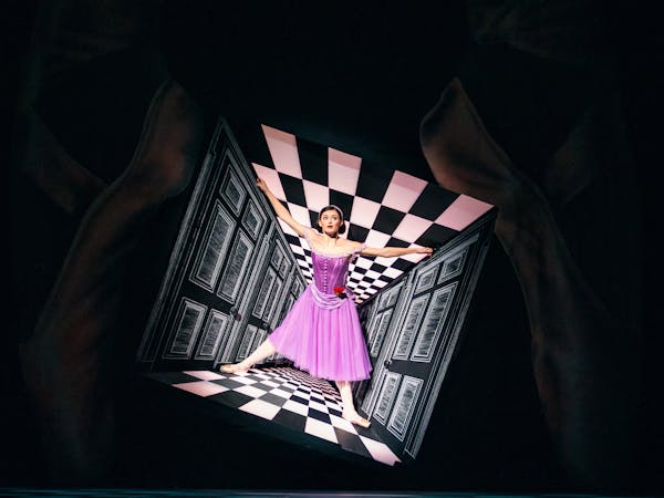 Anna Rose O’Sullivan, portraying Alice in Christopher Wheeldon’s Alice’s Adventures in Wonderland, stands at the centre of a surreal checkered corridor that appears to narrow around her, creating a striking optical illusion. She wears a purple dress and sports a sleek black bob, capturing the whimsical and disorienting essence of Wonderland.