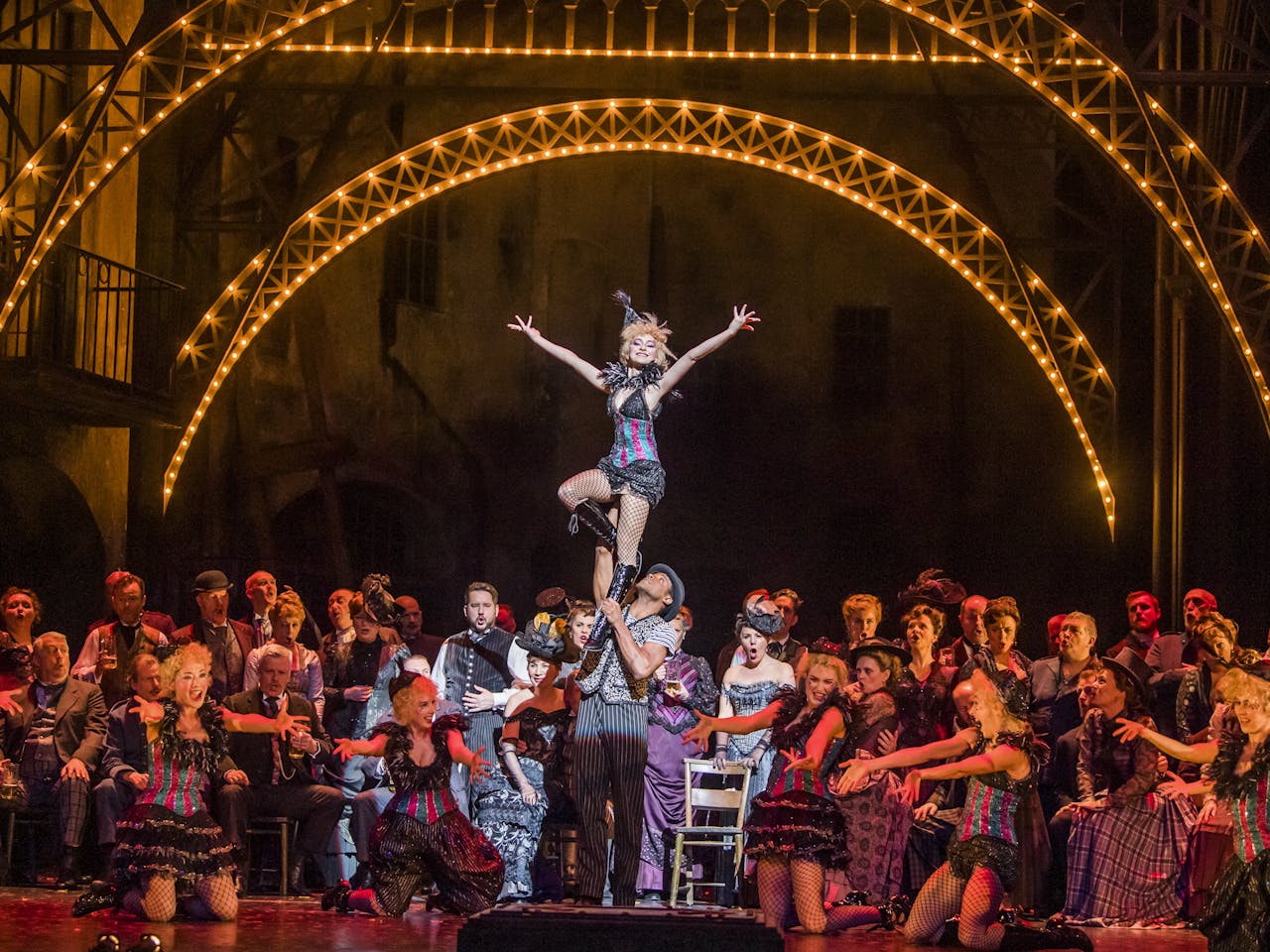 A vibrant and crowded dance scene from the opera Faust, featuring a large ensemble on the Royal Opera House main stage surrounding two lead dancers. The male dancer lifts the smiling female dancer, who stands with her arms outstretched in a joyful pose. The setting evokes a lively 1950s cabaret, with bright stage lights and performers dressed in vintage-style costumes, creating an atmosphere that is both inviting and grand.