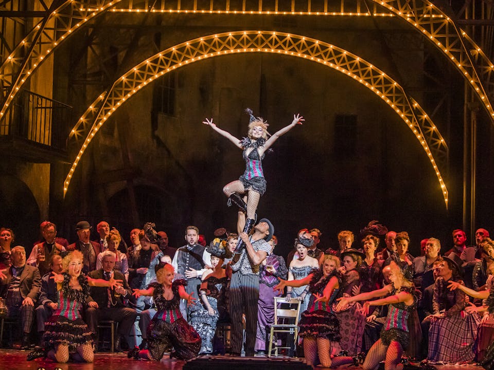 A vibrant and crowded dance scene from the opera Faust, featuring a large ensemble on the Royal Opera House main stage surrounding two lead dancers. The male dancer lifts the smiling female dancer, who stands with her arms outstretched in a joyful pose. The setting evokes a lively 1950s cabaret, with bright stage lights and performers dressed in vintage-style costumes, creating an atmosphere that is both inviting and grand. A vibrant and crowded dance scene from the opera Faust, featuring a large ensemble on the Royal Opera House main stage surrounding two lead dancers. The male dancer lifts the smiling female dancer, who stands with her arms outstretched in a joyful pose. The setting evokes a lively 1950s cabaret, with bright stage lights and performers dressed in vintage-style costumes, creating an atmosphere that is both inviting and grand.