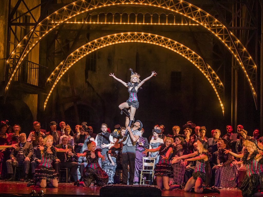 A vibrant and crowded dance scene from the opera Faust, featuring a large ensemble on the Royal Opera House main stage surrounding two lead dancers. The male dancer lifts the smiling female dancer, who stands with her arms outstretched in a joyful pose. The setting evokes a lively 1950s cabaret, with bright stage lights and performers dressed in vintage-style costumes, creating an atmosphere that is both inviting and grand.