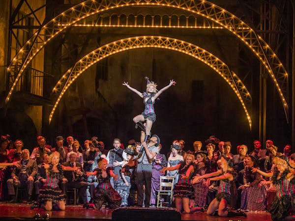 A vibrant and crowded dance scene from the opera Faust, featuring a large ensemble on the Royal Opera House main stage surrounding two lead dancers. The male dancer lifts the smiling female dancer, who stands with her arms outstretched in a joyful pose. The setting evokes a lively 1950s cabaret, with bright stage lights and performers dressed in vintage-style costumes, creating an atmosphere that is both inviting and grand.