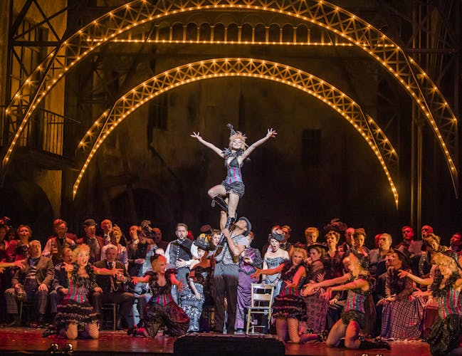 A vibrant and crowded dance scene from the opera Faust, featuring a large ensemble on the Royal Opera House main stage surrounding two lead dancers. The male dancer lifts the smiling female dancer, who stands with her arms outstretched in a joyful pose. The setting evokes a lively 1950s cabaret, with bright stage lights and performers dressed in vintage-style costumes, creating an atmosphere that is both inviting and grand.