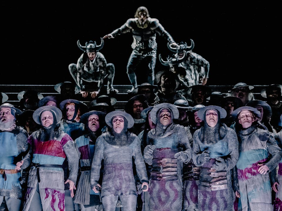 A dramatic scene from Adele Thomas’s Il trovatore, depicting a group of around two dozen men in the centre of the frame, dressed in medieval-style clothing and armor, appearing confused and alarmed. In the background, a blurred, looming figure accompanied by two horned soldiers seems poised to descend upon them, intensifying the sense of impending conflict.