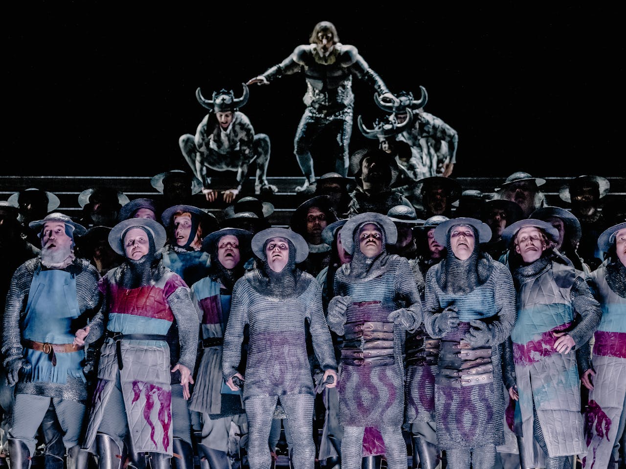 A dramatic scene from Adele Thomas’s Il trovatore, depicting a group of around two dozen men in the centre of the frame, dressed in medieval-style clothing and armor, appearing confused and alarmed. In the background, a blurred, looming figure accompanied by two horned soldiers seems poised to descend upon them, intensifying the sense of impending conflict.