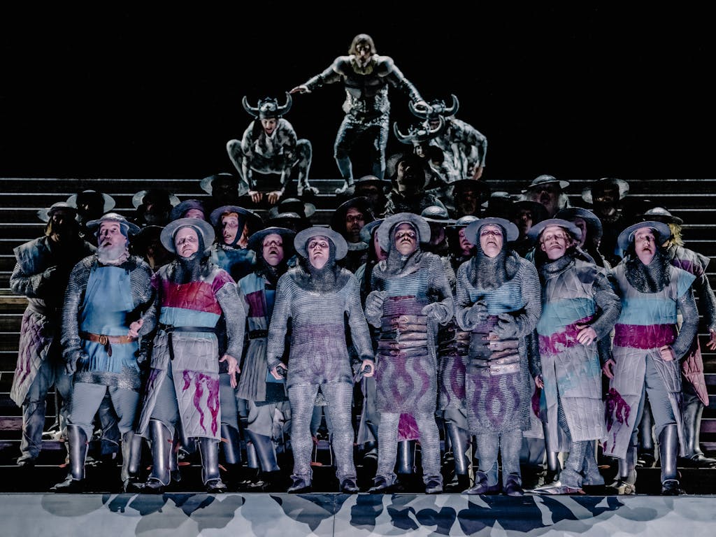 A dramatic scene from Adele Thomas’s Il trovatore, depicting a group of around two dozen men in the centre of the frame, dressed in medieval-style clothing and armor, appearing confused and alarmed. In the background, a blurred, looming figure accompanied by two horned soldiers seems poised to descend upon them, intensifying the sense of impending conflict.