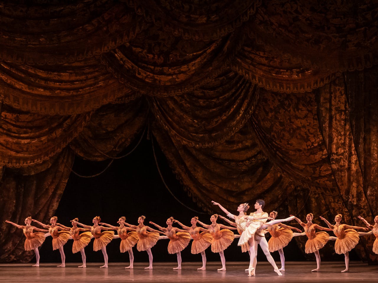 A group of Royal Ballet School dancers on the main stage of the Royal Opera House in synchronised formation, all wearing matching gold tutus, pose in a classic arabesque. The stage is framed by grand, ornate curtains in rich, warm tones, highlighting the elegance and grandeur of the performance.