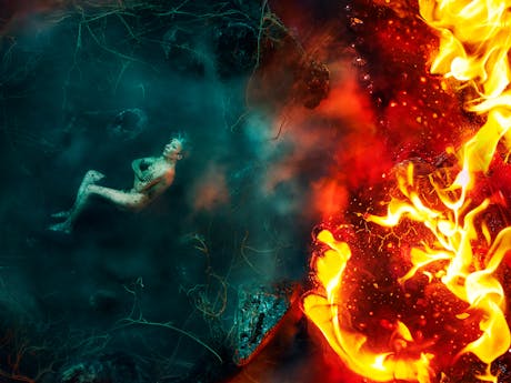 A surreal image divided into two contrasting halves - on the left, a nude figure representing Erda in Barrie Kosky's production of Wagner's Siegfried lies on the ground. She is covered in soil and curled in a fetal position in a misty, dark teal forest environment with scattered rocks and tangled roots; on the right, intense orange flames rage, consuming the space with bright fire and embers, creating a stark contrast between calm and chaos.