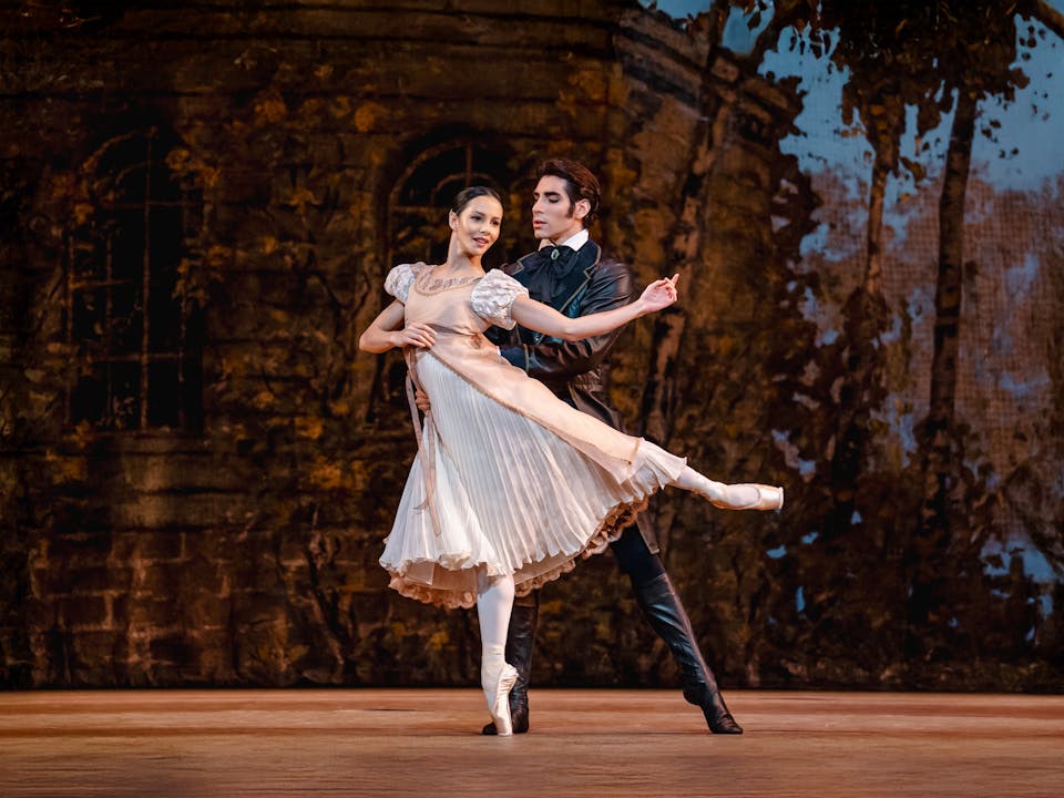 Cesar Corrales and Francesca Hayward perform as Onegin and Tatiana on the Royal Opera House stage. Tatiana, wearing a vintage-style cream dress with puffed sleeves and pointe shoes, balances en pointe with one leg extended behind her. Onegin, dressed in a dark, elegant period costume with knee-high boots, supports her from behind. The backdrop features a shadowy, autumnal forest scene with an old stone building, enhancing the romantic and dramatic atmosphere.