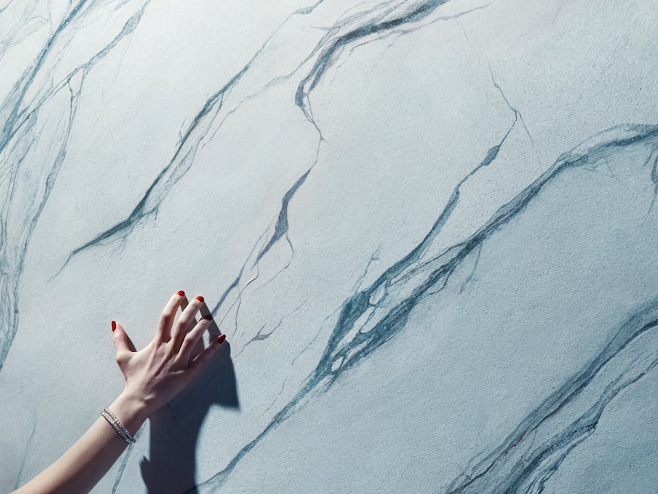 A pale hand with red-painted nails and a silver bracelet reaches towards a marble wall with dark veins running through it. The shadow of the hand is cast on the wall, adding a dramatic effect. The hand belongs to the character playing Tosca in Oliver Mears’s production of Tosca.