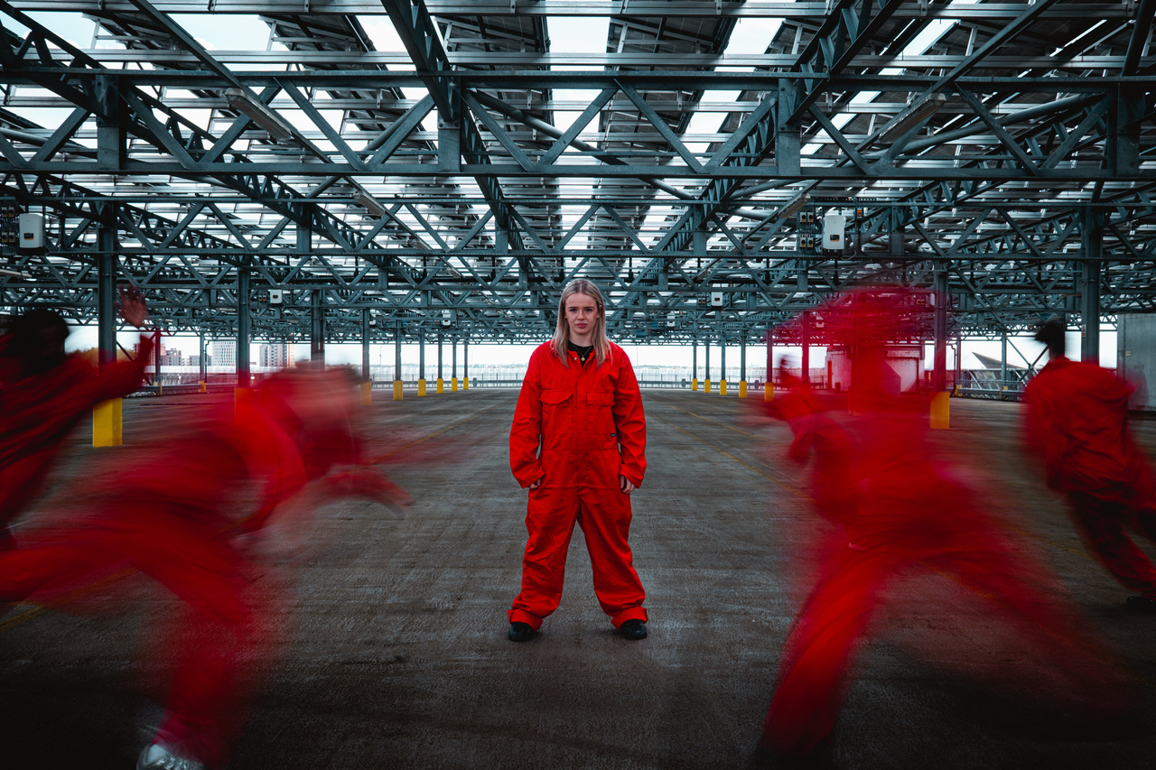 A person stands in the centre of an industrial car park scene wearing an orange boiler suit. Her legs stand apart, her arms hang down by her sides and she looks directly ahead into the camera. In the foreground, several figures also dressed in orange boiler suits rush towards the central figure, their bodies are blurred by motion. 