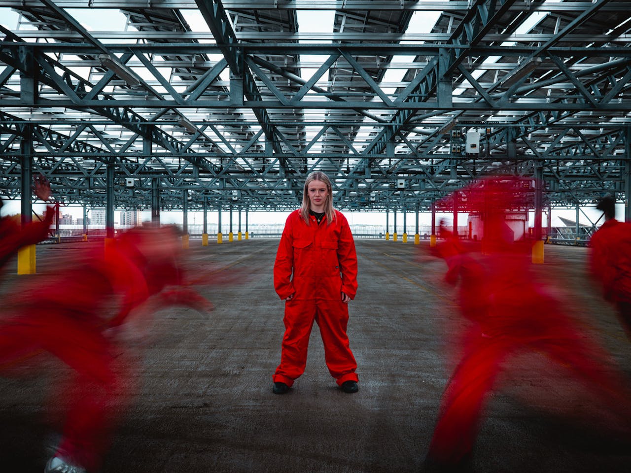A person stands in the centre of an industrial car park scene wearing an orange boiler suit. Her legs stand apart, her arms hang down by her sides and she looks directly ahead into the camera. In the foreground, several figures also dressed in orange boiler suits rush towards the central figure, their bodies are blurred by motion.