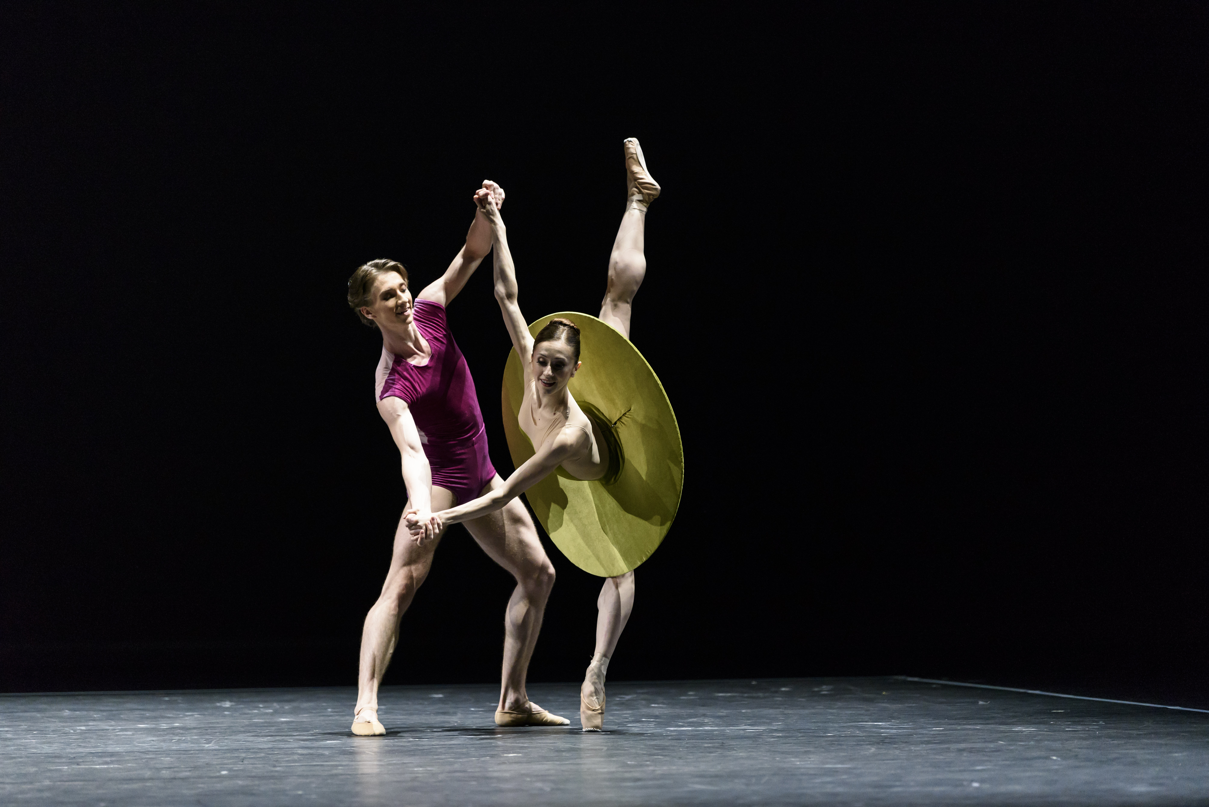 A man, dressed in purple, dances with a woman in a green modern tutu, her leg outstretched behind her. 