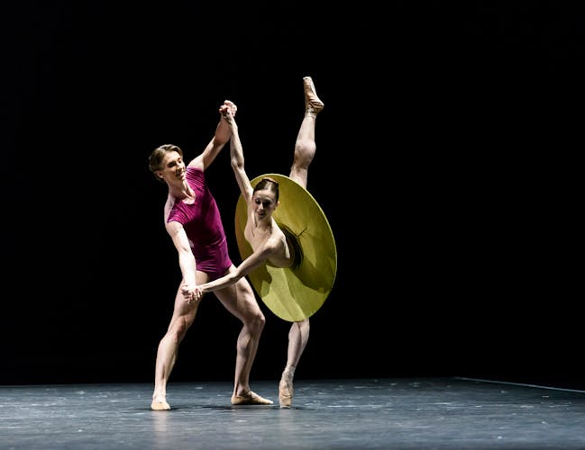 A man, dressed in purple, dances with a woman in a green modern tutu, her leg outstretched behind her.