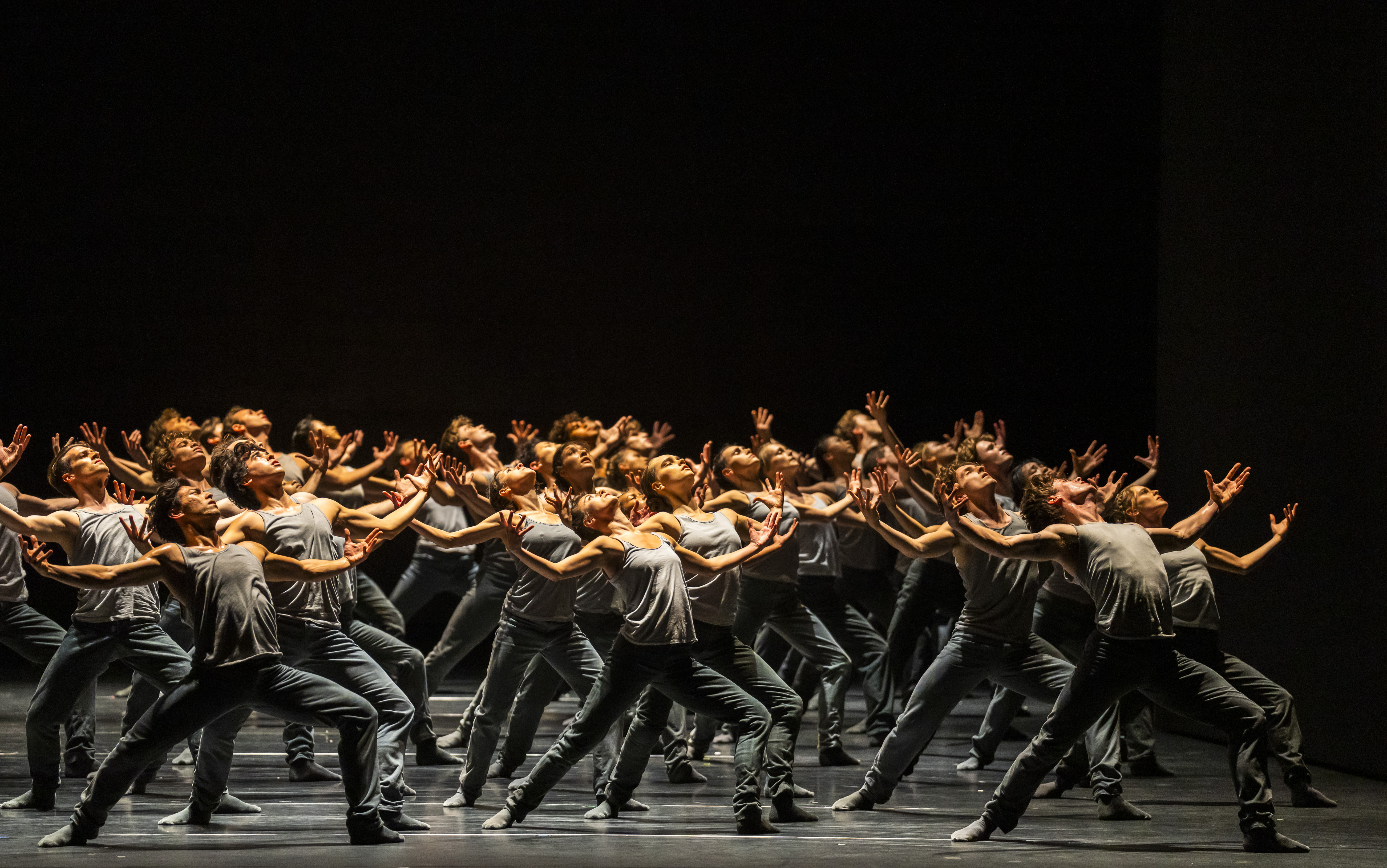 A group of dancers, dressed in grey, stand with their arms outstretched and upper bodies leaning backwards, as if towards the sky.