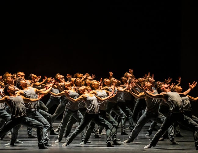 A group of dancers, dressed in grey, stand with their arms outstretched and upper bodies leaning backwards, as if towards the sky.