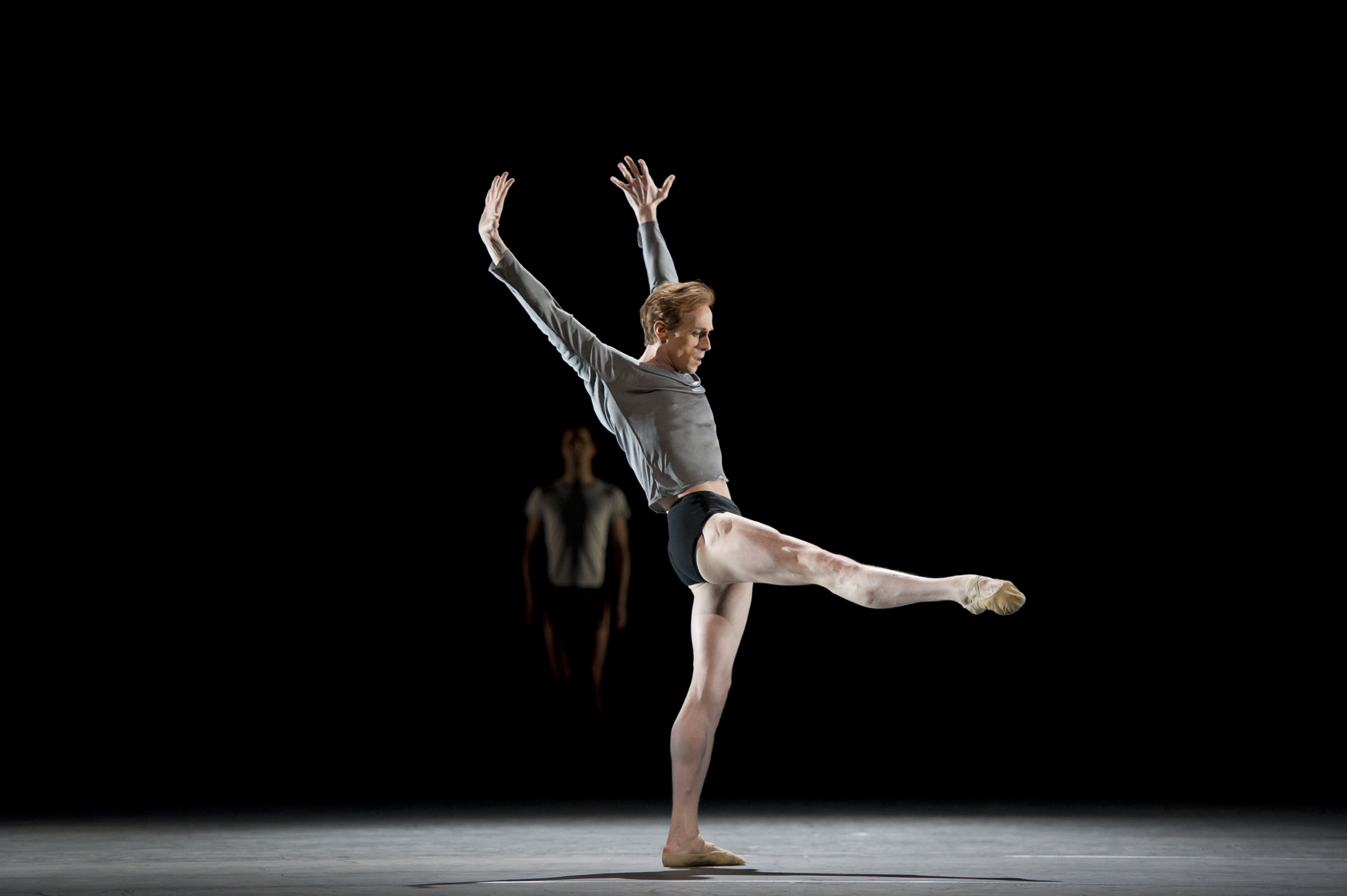 A man stands on one leg, his other leg extended to the front and his two arms stretched above and slightly behind him. 