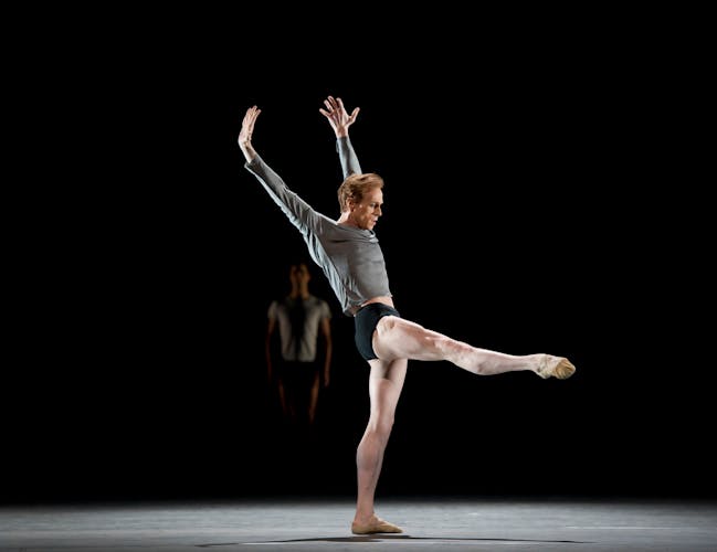 A man stands on one leg, his other leg extended to the front and his two arms stretched above and slightly behind him.