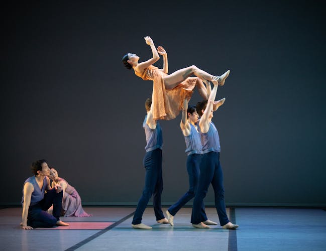 Three men carry a woman above them. Her body is almost horizontal, her legs are bent and her feet are flexed, while her arms are bent. Another man crouches at the corner, watching them.