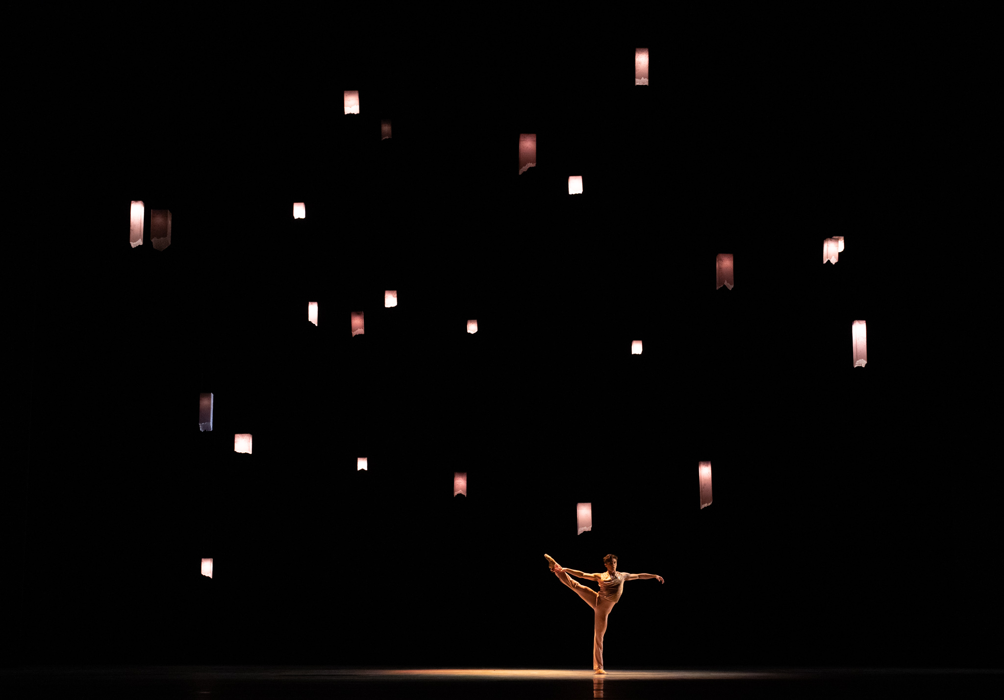 A man dances, solitary, under some lanterns. His arms reach to the side of his body, while his right legs is held outstretched to his side. 